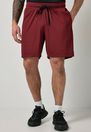 Mand iført mørkrøde sportsshorts med sort snøre, hvide sokker og sorte løbesko, stående mod en ensfarvet lys baggrund.
