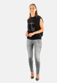 Chaleco negro sin mangas con un logo brillante, combinado con jeans skinny gris claro y tacones negros, sobre un fondo blanco liso.