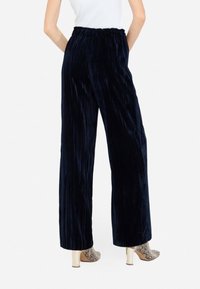 Pantaloni ampi a pieghe di velluto blu navy; vita elastica; abbinati a scarpe con tacco a fantasia, visti da dietro.