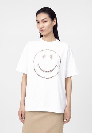 Femme aux cheveux foncés et mouillés porte un t-shirt blanc oversize avec un motif de visage souriant perforé et une jupe beige sur fond uni.