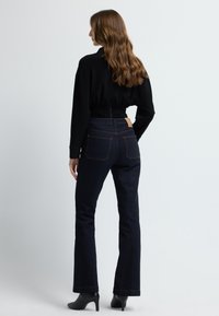 Musta pitkähihaine paita, jossa on solmittu selkä, yhdistettynä tummansinisille leveälahkeisille farkuille, joissa on näkyvää ompelua ja vaaleanruskea nahkalappu takana.