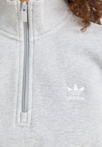 Szary polarowy sweter z wysokim kołnierzem i zamkiem na ćwierć długości. Posiada małe białe logo Adidasa na piersi. Miękka tekstura.