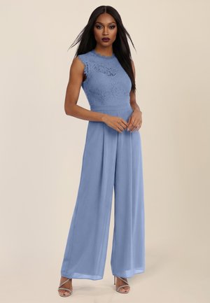 Frau, die in einem ärmellosen, himmelblauen Spitzen-Jumpsuit mit weitem Bein und silbernen Riemchen-High Heels vor einem schlichten beigen Hintergrund steht.