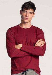 Bordeaux langwerpige t-shirt met ronde hals, gemaakt van gladde stof. Model poseert met de armen gekruist, terwijl hij grijze broek met een trekkoord draagt.