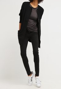 Lång svart kofta med knappstängning, kombinerad med en mörkgrå t-shirt och smala svarta jeans. Skorna är vita sneakers med svarta detaljer.