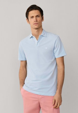 Man in een lichtblauw poloshirt met korte mouwen en roze broek, staand tegen een eenvoudige neutrale achtergrond.