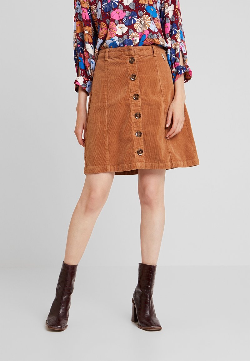 Femme portant une jupe en velours côtelé marron à boutons sur le devant, un blouse à manches longues à fleurs, et des bottines à talons marron foncé se tenant sur un fond uni.