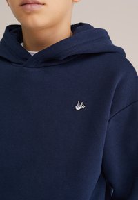 Tumed sinine kapuutsiga särk, mis on valmistatud puuvillast ja millel on ees suur tasku. Vasakul rinnal on väike valge linnu logo.