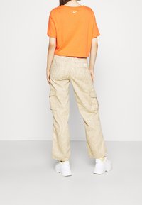 Orange kortärmad t-shirt med rund hals och en vit logotyp; beige cargobyxor med spiral mönster och sidofickor, i kombination med vita sneakers.