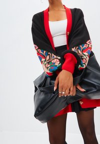 Cardigan nero con accenti rossi, caratterizzato da ricami floreali colorati sulle maniche. La texture è a maglia; i bottoni sono visibili. Borsa nera tenuta in mano.