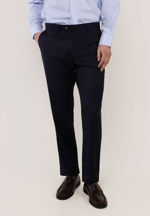 Hombre con pantalones de vestir azul marino, camisa azul claro abotonada y mocasines de cuero marrón oscuro, de pie con una mano en el bolsillo.