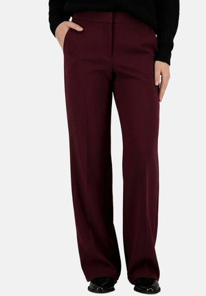 Straight-leg burgundy pants worn with a black sweater, one hand in pocket, and black shoes visible on a white background.