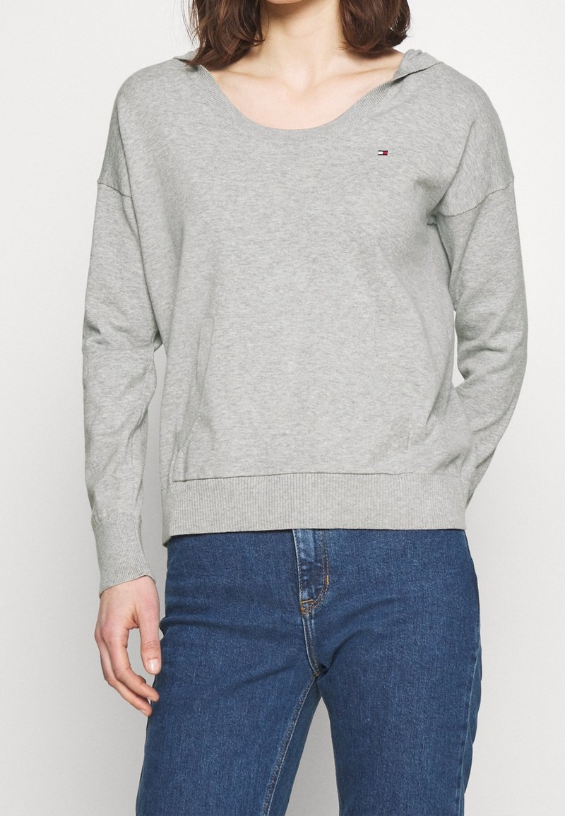 Femme portant un sweat-shirt gris clair à manches longues avec un petit logo sur la poitrine et un jean en denim bleu.