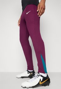 Nike sporta legingi tumši purpursarkanā krāsā ar ribotu tekstūru, ar teļa akcenta elementu un nelielu baltu logo priekšpusē. Parādīti baltie sporta apavi.
