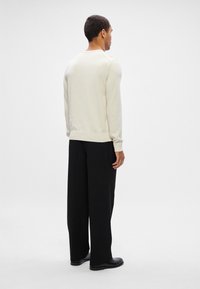 Ein Mann steht mit dem Rücken zur Kamera und trägt einen cremfarbenen Langarmpullover, schwarze weite Hosen und schwarze Schuhe vor einem schlichten weißen Hintergrund.