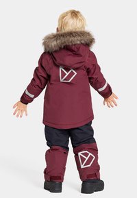 Mörkröd parkas med fuskpäls på huven, reflekterande detaljer och geometrisk logotyp på baksidan. Kombinerad med marinblå snöbyxor och svarta stövlar.