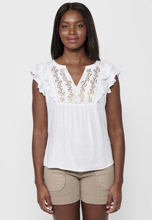Blouse blanche avec des manches capes à volants, ornée de perles décoratives dorées et noires sur le devant, et d'un décolleté en V. Tissu texturé.