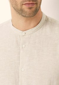 Camicia button-up beige chiaro con scollatura rotonda, caratterizzata da un tessuto testurizzato e due bottoni bianchi al colletto.