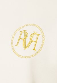 Valkoinen kangas, jossa on pyöreä kultalankakirjailtu logo, joka esittää toisiinsa kietoutuneita kirjaimia "R" ja "A" ympyräiseen ketjumuotoon.