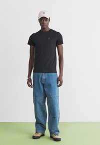 Tricou negru cu mânecă scurtă, prevăzut cu o buzunar pe piept, asortat cu blugi denim deschiși, cu picior larg și cusături panel. Căciulă de baseball bej și pantofi deschiși.