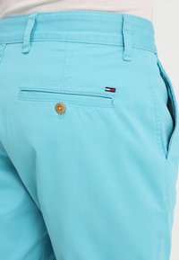 Tyrkysové šortky vyrobené z bavlny. Mají zadní kapsu s dřevěným knoflíkem a malou nášivku v červené, bílé a modré barvě na boku.