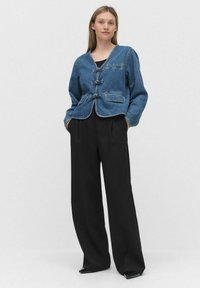 Veste en denim avec un décolleté en V, fermeture à boutons sur le devant et deux poches avant, associée à un pantalon noir taille haute et des chaussures noires à bout pointu.