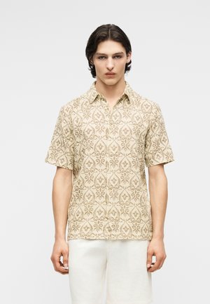 Νεαρός άνδρας με σκούρα μαλλιά, που φοράει μπεζ κοντομάνικο πουκάμισο με επαναλαμβανόμενο περίτεχνο σχέδιο και λευκά σορτς, μπροστά σε απλό φόντο.