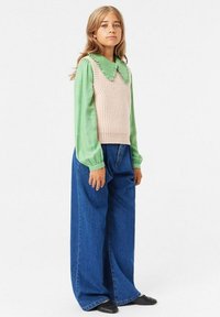 Blusa a maniche lunghe verde chiaro con colletto a scallop abbinata a un gilet beige a coste, pantaloni in denim blu larghi e ballerine nere.