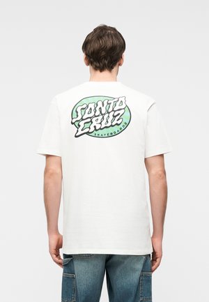 Άνδρας που φοράει λευκό μπλουζάκι με πράσινο οβάλ λογότυπο «Santa Cruz Skateboards» στην πλάτη και μπλε τζιν cargo, όρθιος με την πλάτη προς τα εμπρός.