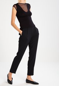 Top negro ajustado con escote de malla y mangas cortas, combinado con pantalones negros de corte clásico con un ajuste relajado y pliegues en la parte frontal.