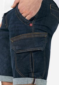 Dark denim shorts with rolled cuffs, featuring yellow stitching and a side pocket with a flap, accented by a small red logo.
