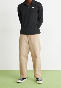 Svart långärmad polo-tröja med liten vit logotyp, kombinerad med beige, löst sittande byxor och svarta sneakers med vita sulor.