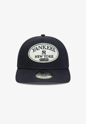 Črna kapa z ovalno belo zakrpano. Besedilo pravi "YANKEES NEW YORK EST. 1903." Ima ukrivljen vizir in nastavljiv hrbet.