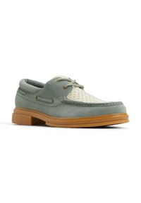 Zapato de barco verde de ante con parte superior entrelazada en beige, puntera redondeada y suela de goma. Presenta cierre con cordones y detalles de costura en contraste.