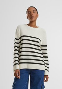 Maglione crema con strisce orizzontali nere, texture a costine, colletto rotondo e maniche lunghe. Abbinato a pantaloni in denim blu a gamba larga.