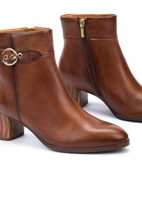 Botas de cuero marrón hasta el tobillo con punta afilada, cremallera lateral y tacón bloque redondo con textura de madera. Presenta un acento de hardware circular dorado.