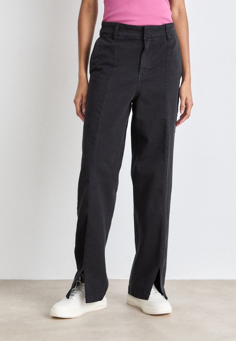 Pantaloni neri a gamba larga con orli spaccati, realizzati in cotone, dotati di cerniera frontale e tasche. Indossati con sneakers bianche.