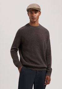 Brun ribbet genser med rund hals og lange ermer, parret med mørke bukser. Modellen har på seg en lys grå flat caps.