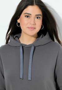 Sudadera gris con capucha y cordón en un tono azul contrastante, con puntas de metal plateadas. Textura de tela suave, corte relajado.