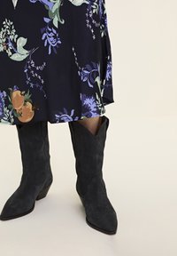 Cizme de cowboy din suede negru, cu vârfuri ascuțite și laturi unghiulare, purtate cu o rochie cu model floral în tonuri închise, incluzând verde și mov.