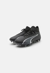Scarpe da calcio nere con una finitura testurizzata, dotate di un colletto alto, lacci neri e una striscia bianca a lato. Tacchetti trasparenti sulla suola.