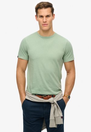 Hellgrünes Baumwoll-T-Shirt mit Rundhalsausschnitt und kurzen Ärmeln. Kombiniert mit marineblauen Hosen, grauem Pullover um die Taille gebunden, braunem Gürtel und Uhr.