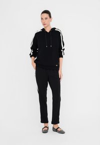 Mujer vestida con sudadera negra y pantalones con rayas blancas, de pie con una mano en el bolsillo y zapatos planos grises sobre fondo blanco.