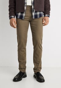 Άνδρας που φοράει παντελόνι σε στενή γραμμή σε ελιά πράσινο χρώμα, μαύρα μοκασίνια, με στρώσεις από μπλε καρό πουκάμισο και σκούρο καφέ μπουφάν με φερμουάρ.