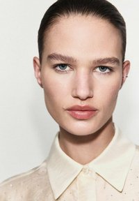 Portrait of a person with smooth skin, light brown hair, wearing a beige collared shirt with subtle embellishments, featuring blue eyes and natural makeup.