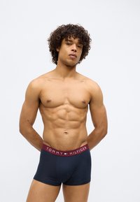 Mišićavi muškarac s kovrčavom kosom stoji nasuprot jednostavne pozadine, odjeven u mornarsko plave Tommy Hilfiger bokserice s burgundskom elastičnom trakom.