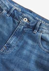 Sininen farkkuinen skinny fit -housut, joissa on nappikiinnitys, etutaskut, vyölenkit ja sisävyötäröllä teksti "NEXT" ja "SKINNY."