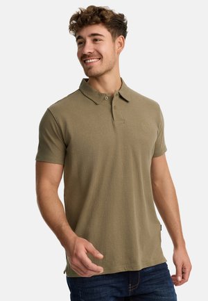 Camiseta polo verde oliva en mezcla de algodón, de mangas cortas, con tapeta de dos botones, pequeño logo en el pecho, combinada con jeans azul oscuro.