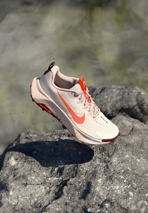 Lyse beige Nike løbesko med orange swoosh og detaljer svævende over en struktureret grå sten udendørs.
