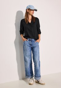 Maglietta nera a maniche lunghe, cappellino azzurro chiaro, jeans a gamba larga blu sbiaditi e sneakers bianche con dettagli neri, in piedi di fronte a uno sfondo chiaro.